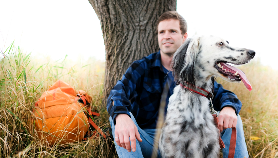 Un bărbat stătea în fața unui copac cu rucsacul portocaliu pe iarbă și câinele lui stătea în fața lui în concediu